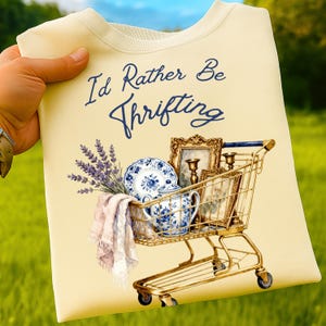 May include: Cream-colored sweatshirt with the text "I'd Rather Be Thrifting." The design includes a gold shopping cart filled with vintage items: a blue and white floral teacup and plate, framed pictures, and lavender sprigs.