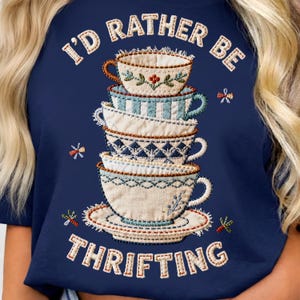 Może przedstawiać: Biały ceramiczny kubek z czarnym uchwytem i napisem "But First, Coffee" pogrubioną, czarną czcionką. Kubek jest umieszczony na rozmytym tle, sugerującym przytulne otoczenie.