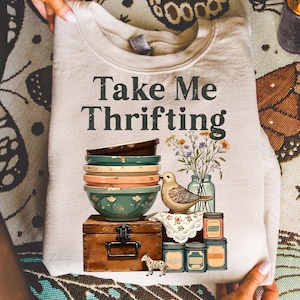 May include: Cream-colored sweatshirt with the text "Take Me Thrifting." The graphic features a stack of bowls, a bird, flowers in a jar, and a small chest. A small decorative dog and jars are also included in the design.