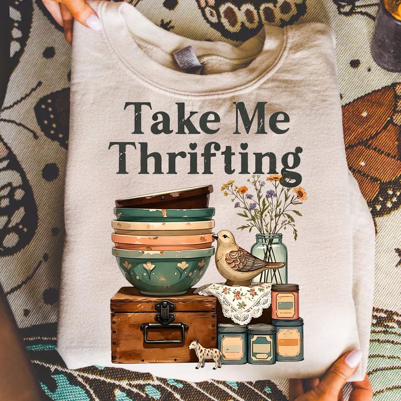 May include: Cream-colored sweatshirt with the text "Take Me Thrifting." The graphic features a stack of bowls, a bird, flowers in a jar, and a small chest. A small decorative dog and jars are also included in the design.