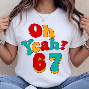 May include: White t-shirt with the text "Oh Yeah! 67" in a retro font. The text is in red, yellow, and teal colors. The shirt has short sleeves and a round neckline.