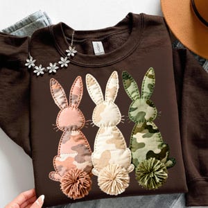 May include: A brown sweatshirt featuring three appliqued bunny rabbits with camouflage patterns in pink, cream, and green. Each bunny has a matching pom-pom tail. A floral necklace and a hat are visible in the background.