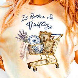 May include: Cream-colored t-shirt with the text "I'd Rather Be Thrifting" in navy blue script. A gold shopping cart is filled with vintage items, including a blue and white floral teacup and plate, framed pictures, and lavender.