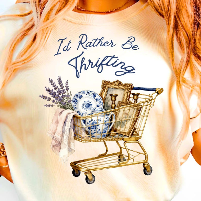May include: Cream-colored t-shirt with the text "I'd Rather Be Thrifting" in navy blue script. A gold shopping cart is filled with vintage items, including a blue and white floral teacup and plate, framed pictures, and lavender.