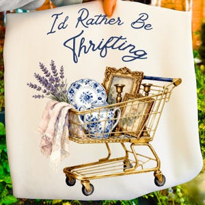 May include: A watercolor illustration features a gold shopping cart filled with vintage items. The cart holds blue and white china, framed pictures, and candles. The text "I'd Rather Be Thrifting" is written in navy blue script.