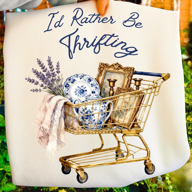 May include: A watercolor illustration features a gold shopping cart filled with vintage items. The cart holds blue and white china, framed pictures, and candles. The text "I'd Rather Be Thrifting" is written in navy blue script.