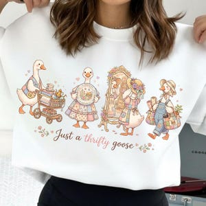 以下が含まれることがあります： ヴィンテージの服を着たガチョウの気まぐれなイラストが特徴の白いスウェットシャツ。「Just a thrifty goose」のテキストがスクリプトフォントで表示され、さまざまなアクセサリーや小道具が描かれています。