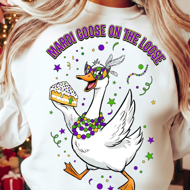 May include: White sweatshirt featuring a cartoon goose wearing a Mardi Gras mask and beads, holding a slice of cake. The text "MARDI GOOSE ON THE LOOSE" arches above the goose. The design includes stars, confetti, and swirls in purple, green, and yellow.