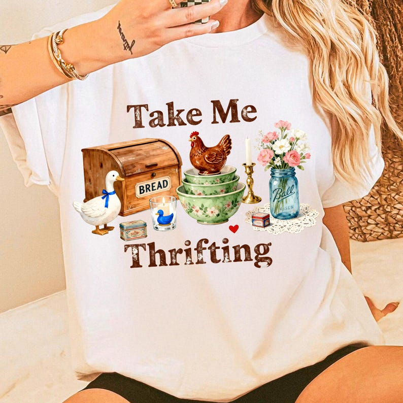 May include: White t-shirt with the text "Take Me Thrifting" and various illustrations. The illustrations include a wooden bread box, a duck with a blue ribbon, a chicken, bowls, a candle, a jar of flowers, and other small items.