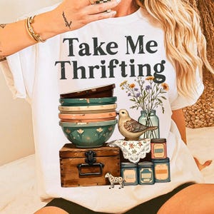 May include: White t-shirt with the text "Take Me Thrifting" in black. The shirt features a stack of colorful bowls, a wooden chest, a bird, flowers in a jar, and decorative tins. A small decorative dog is also present.