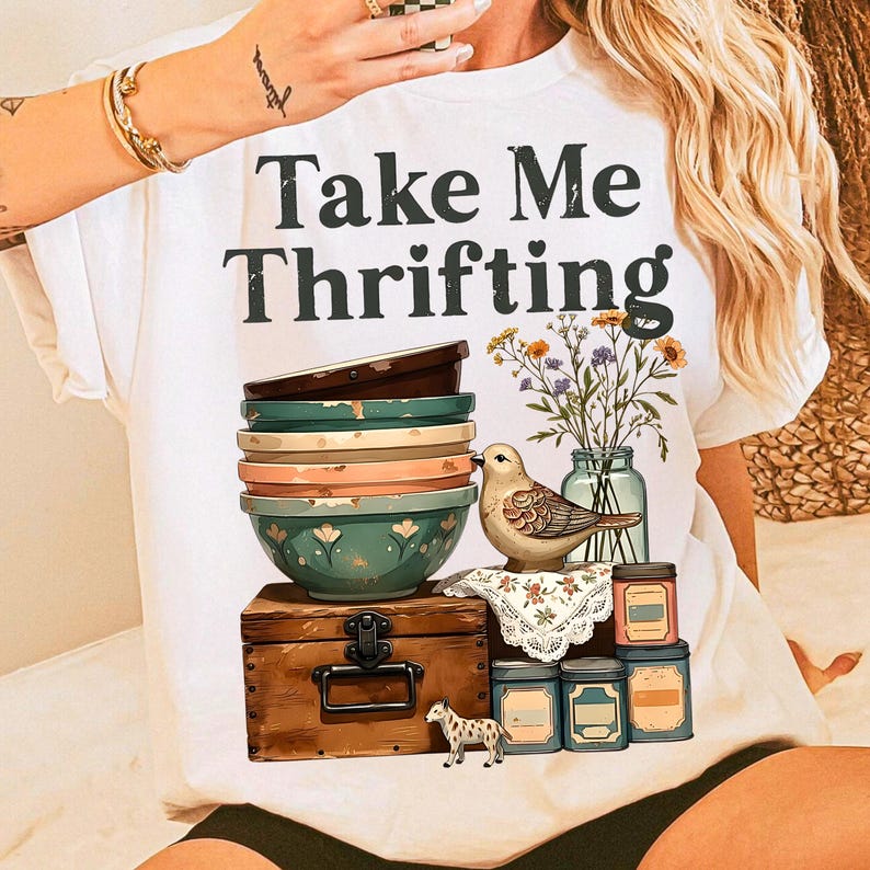 May include: White t-shirt with the text "Take Me Thrifting" in black. The shirt features a stack of colorful bowls, a wooden chest, a bird, flowers in a jar, and decorative tins. A small decorative dog is also present.