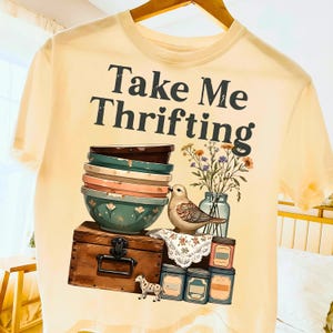 May include: Cream-colored t-shirt with the text "Take Me Thrifting" in black. The graphic features a stack of bowls, a bird, flowers in a jar, a wooden chest, and small jars. A small decorative horse is also present.