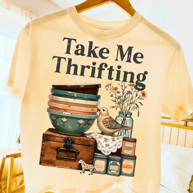 May include: Cream-colored t-shirt with the text "Take Me Thrifting" in black. The graphic features a stack of bowls, a bird, flowers in a jar, a wooden chest, and small jars. A small decorative horse is also present.