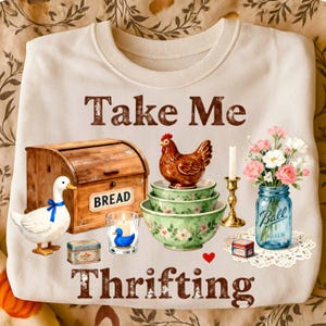 May include: Cream-colored sweatshirt with the text "Take Me Thrifting." The design includes a wooden bread box, a ceramic duck, a chicken figurine, floral bowls, a candle, and a blue Ball jar filled with flowers.