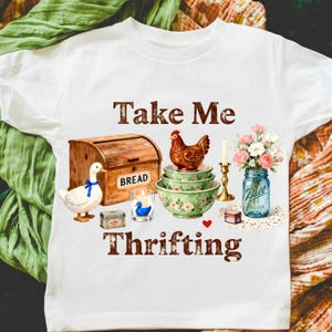 May include: White t-shirt with a graphic featuring a wooden bread box, duck, chicken, bowls, candle, flowers in a jar, and the text "Take Me Thrifting". The design has a vintage, farmhouse aesthetic.