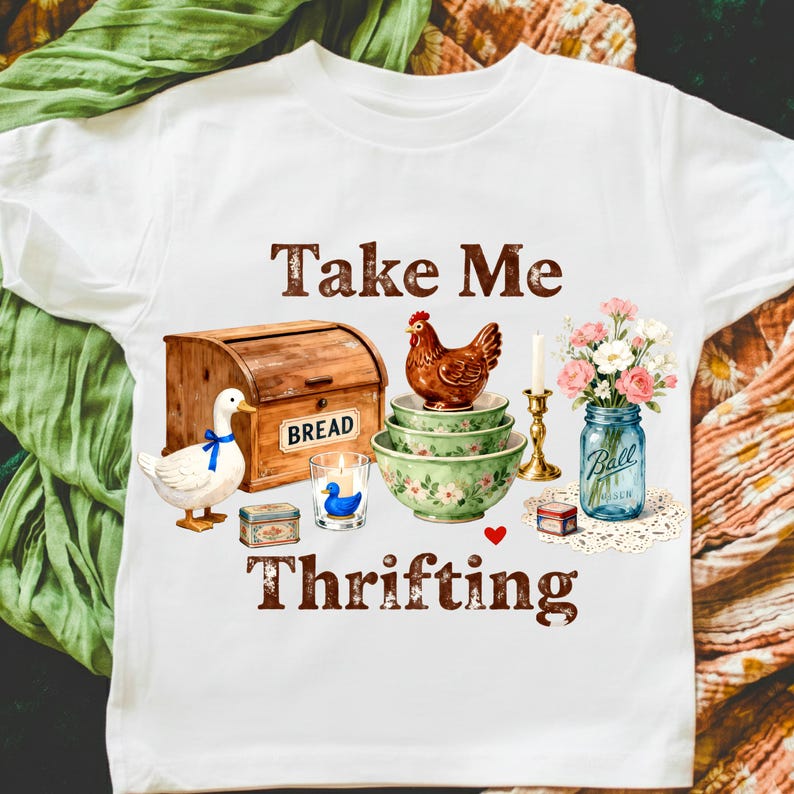 May include: White t-shirt with a graphic featuring a wooden bread box, duck, chicken, bowls, candle, flowers in a jar, and the text "Take Me Thrifting". The design has a vintage, farmhouse aesthetic.