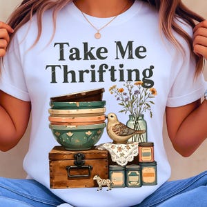 May include: White t-shirt with the text "Take Me Thrifting" above a graphic of stacked bowls, a bird, flowers in a jar, and a wooden chest. The graphic also includes small jars and a decorative cloth.