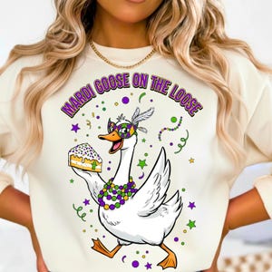 May include: Cream-colored sweatshirt featuring a cartoon goose in a Mardi Gras mask, holding a slice of cake. The goose is adorned with a beaded necklace and a feathered headpiece. The text "MARDI GOOSE ON THE LOOSE" arches above the goose in purple and green.