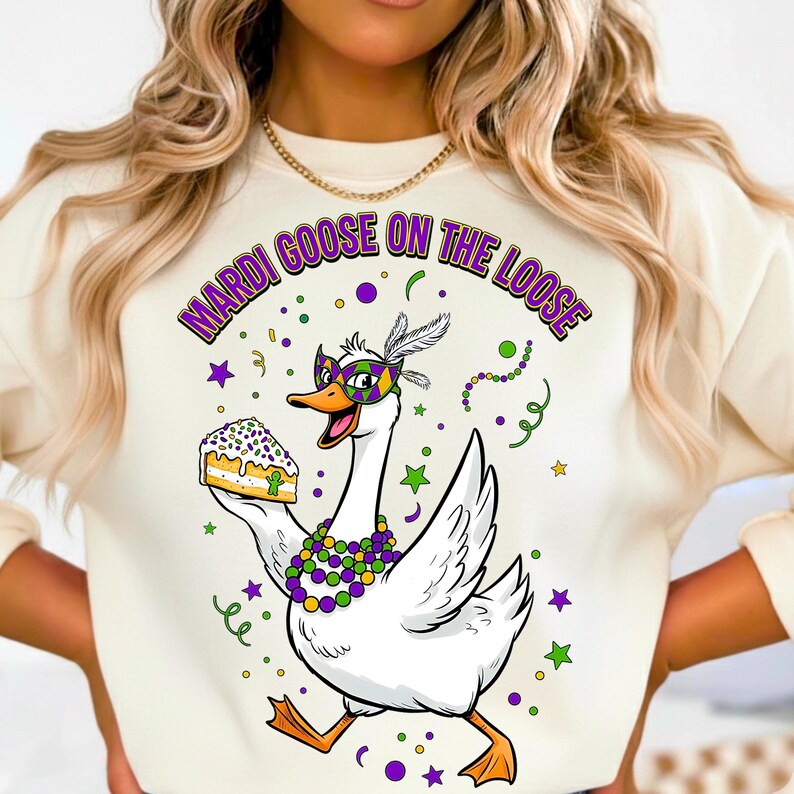 May include: Cream-colored sweatshirt featuring a cartoon goose in a Mardi Gras mask, holding a slice of cake. The goose is adorned with a beaded necklace and a feathered headpiece. The text "MARDI GOOSE ON THE LOOSE" arches above the goose in purple and green.