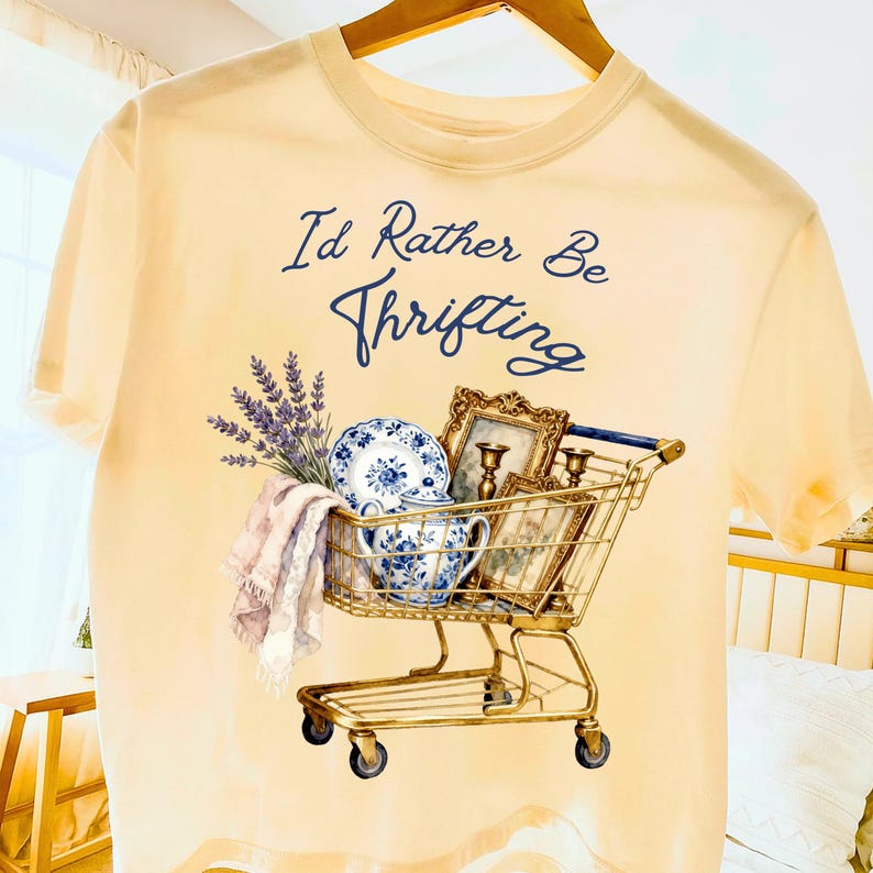 May include: A light yellow t-shirt with the text "I'd Rather Be Thrifting" in navy blue script. The shirt features a watercolor design of a shopping cart filled with vintage items, including a blue and white plate, framed pictures, and lavender.