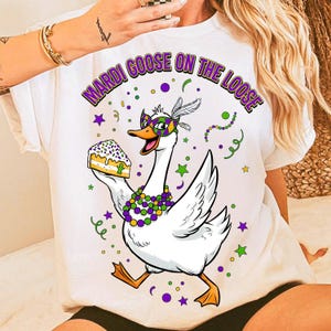 May include: White t-shirt featuring a cartoon goose wearing a mask and holding a slice of cake. The text "MARDI GOOSE ON THE LOOSE" is arched above the goose. The design includes purple, green, and yellow accents, with stars and confetti.