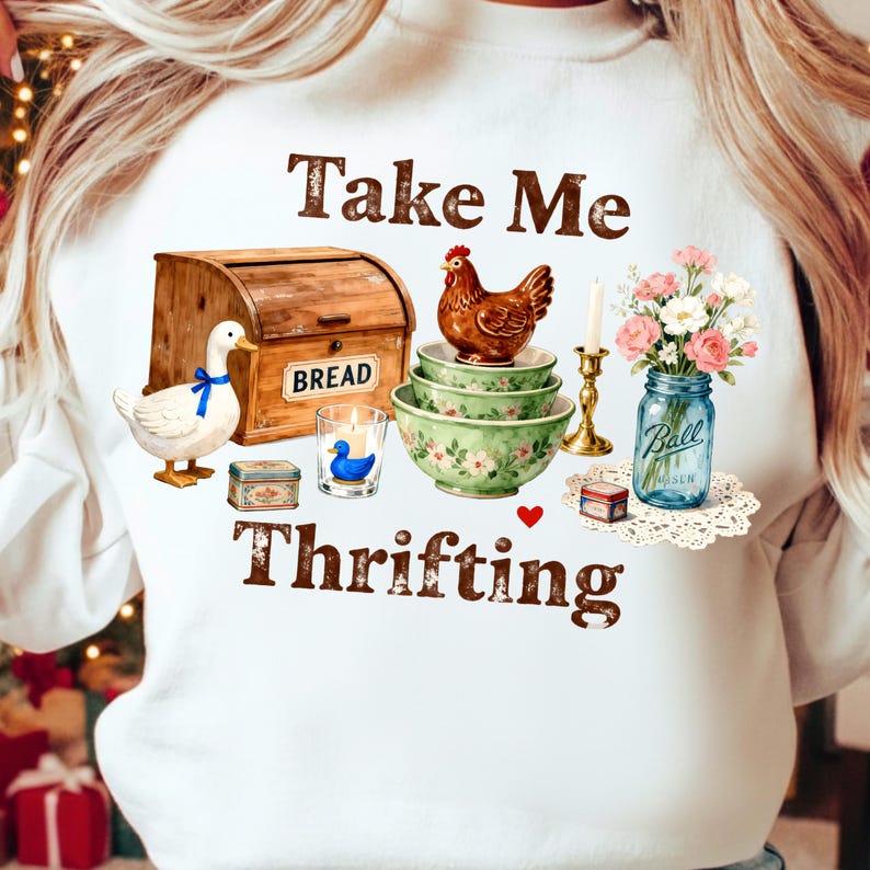 May include: White sweatshirt with a graphic featuring a wooden bread box, a duck, a chicken, floral bowls, a candle, a jar of flowers, and the words "Take Me Thrifting."