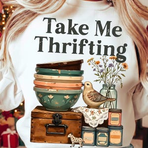 May include: White sweatshirt with the text "Take Me Thrifting" above a graphic of stacked bowls, a bird, a wooden chest, and jars. The bowls are in shades of green, orange, and brown. A small vase of flowers is also present.