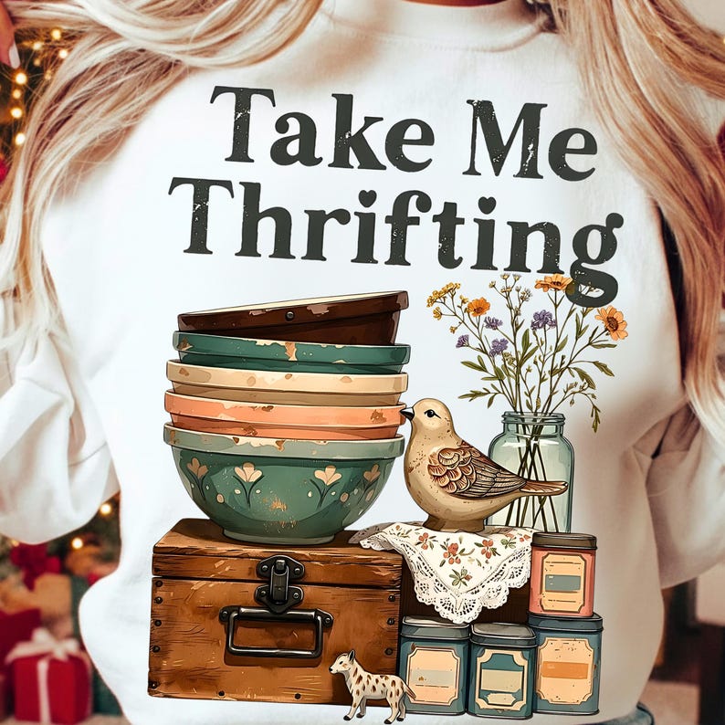 May include: White sweatshirt with the text "Take Me Thrifting" above a graphic of stacked bowls, a bird, a wooden chest, and jars. The bowls are in shades of green, orange, and brown. A small vase of flowers is also present.