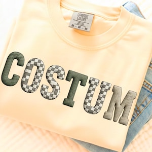 May include: A cream-colored sweatshirt with the word "COSTUM" embroidered in a mix of green, checkered, and gray letters. The sweatshirt is folded, with a pair of blue jeans visible on the side. The tag reads "COMFORT COLORS".