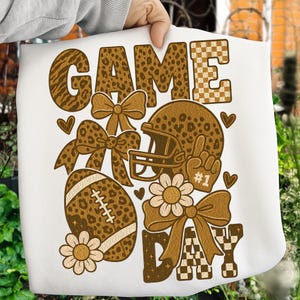 May include: White graphic tee with a football theme. The design features the words "GAME DAY" in brown with leopard print and checkered patterns. Includes a football, helmet, bows, flowers, and a hand gesture.