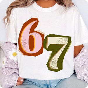 May include: White t-shirt featuring the numbers "6" and "7" in a retro style. The number "6" is pink with a yellow outline and a brown border. The number "7" is off-white with a green border.