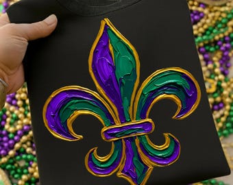 May include: Black sweatshirt featuring a vibrant fleur-de-lis design in purple, green, and gold. The emblem is prominently displayed, with a textured, painted appearance. The sweatshirt is a casual garment, perfect for Mardi Gras celebrations.