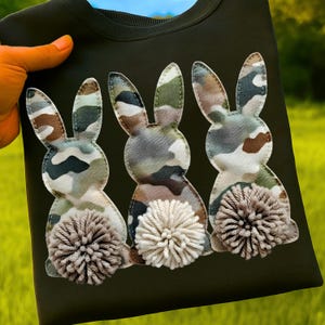 May include: Dark green t-shirt featuring three appliqued bunny rabbits. The bunnies have camouflage patterned bodies and fluffy, cream-colored pom-pom tails. The shirt is held up against a background of green grass and blue sky.