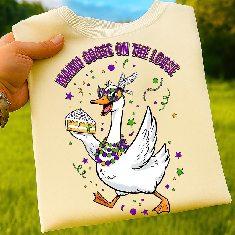 May include: Cream-colored sweatshirt with a graphic of a goose wearing a mask and holding cake. The text "MARDI GOOSE ON THE LOOSE" is arched across the top in purple and green. The goose is decorated with beads and feathers.