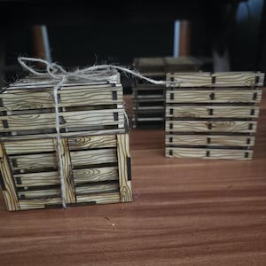 May include: A stack of miniature wooden pallets tied together with thin twine, featuring a light tan color with dark brown wood grain patterns and visible horizontal slats. A single matching miniature wooden pallet stands upright nearby.