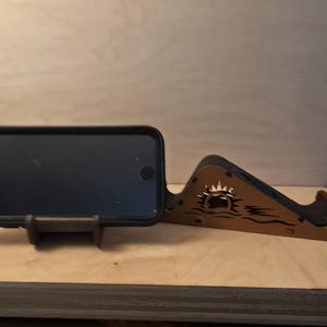 Puede incluir: Un soporte de teléfono de madera con un teléfono negro en una funda. El soporte es marrón con un diseño decorativo de sol y olas. El teléfono descansa sobre un pequeño soporte marrón oscuro.