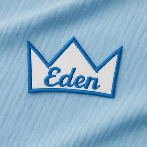 Peut inclure: Un tissu bleu clair avec un écusson brodé blanc et bleu. L'écusson représente une couronne avec le nom "Eden" en écriture bleue. Le tissu a une texture côtelée subtile.