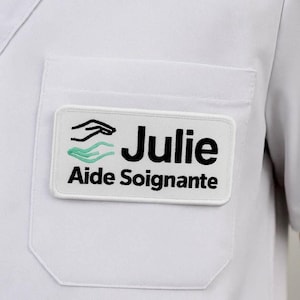 Pode incluir: Uma etiqueta branca com o nome "Julie" em letras pretas e o título "Aide Soignante" abaixo. A etiqueta apresenta um desenho de mão preta e ondas turquesas. A etiqueta está presa a um bolso de camisa branca.