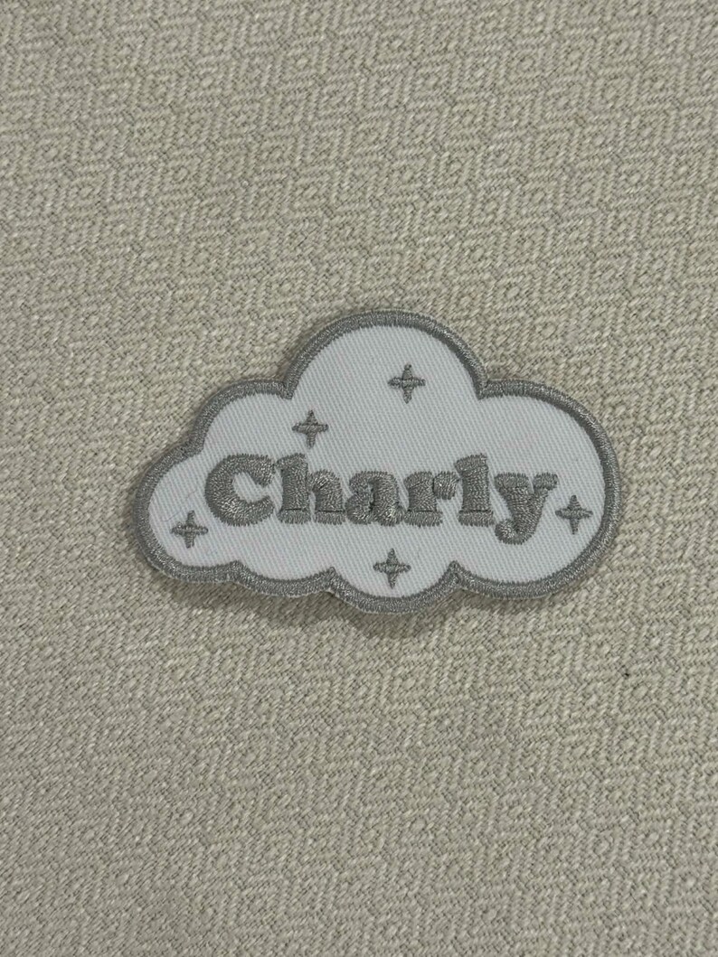 May include: A white cloud-shaped patch with gray embroidered trim and the name "Charly" in gray lettering. Four gray star shapes are embroidered around the name. The patch is on a textured, light beige background.