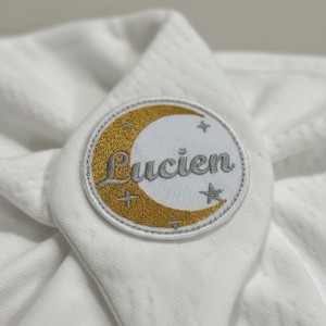Peut inclure: Une couverture blanche pour bébé avec un écusson circulaire brodé du nom "Lucien". L'écusson présente un croissant de lune doré, des étoiles et le nom en fil argenté. La couverture est pliée, créant un aspect texturé.