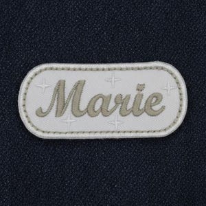 Puede incluir: Un parche ovalado blanco con el nombre "Marie" bordado en escritura marrón claro. El parche tiene un borde gris claro y cuatro pequeños diseños de estrellas. El fondo es una tela azul oscuro texturizada.