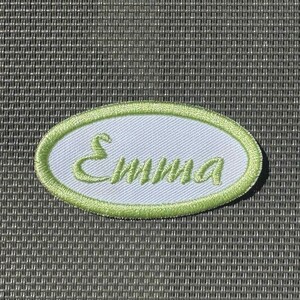 Può includere: Una toppa ricamata ovale con il nome "Emma" in corsivo verde. La toppa ha un centro bianco e un bordo verde chiaro. Lo sfondo è una superficie grigia strutturata.