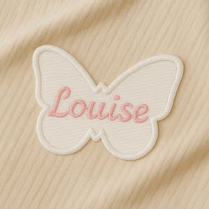 Peut inclure: Un écusson en forme de papillon crème avec le nom "Louise" brodé en rose en écriture cursive. L'écusson a une bordure blanche et est posé sur un fond beige en tissu côtelé.