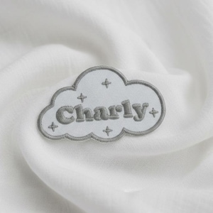 May include: A light gray cloud-shaped patch with the name "Charly" embroidered in a darker gray font. The patch has a gray border and four small cross-shaped designs. The patch is on a white fabric background.