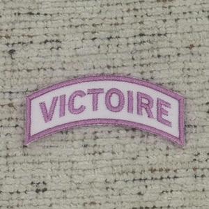 Könnte beinhalten: Ein weißes, gebogenes Abzeichen mit dem Wort "VICTOIRE" in Lavendelfaden gestickt. Das Abzeichen hat einen lavendelfarbenen Rand und ist zum Aufnähen auf Kleidung oder Stoffartikel bestimmt.
