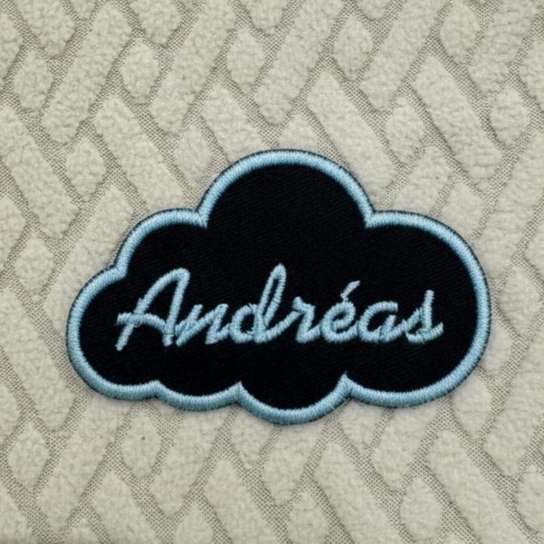 May include: A black cloud-shaped patch with the name "Andreas" embroidered in light blue cursive. The patch has a light blue border and is set against a textured, off-white background.