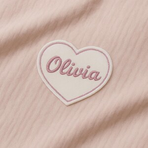 Könnte beinhalten: Ein herzförmiger Aufnäher mit dem Namen "Olivia" in Schreibschrift gestickt. Der Aufnäher hat eine weiße Mitte mit einem rosa Rand und ist vor einem hellrosa Stoffhintergrund platziert.