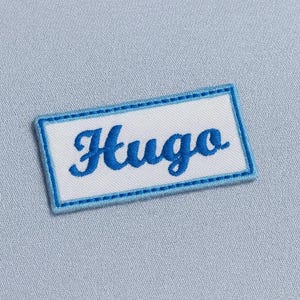 Può includere: Una toppa rettangolare con il nome "Hugo" ricamato in blu. La toppa ha uno sfondo bianco, un bordo azzurro ed è posta su un tessuto azzurro chiaro.