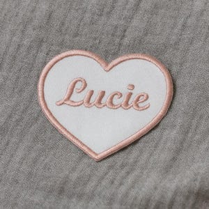 Peut inclure: Un écusson en forme de cœur avec un centre blanc et une bordure rose. Le nom "Lucie" est brodé en rose en écriture cursive à l'intérieur du cœur. L'écusson est posé sur un fond en tissu gris.