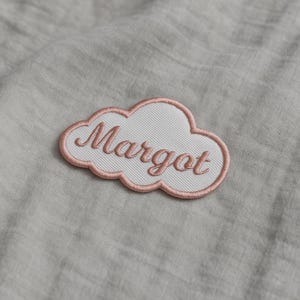 Könnte beinhalten: Ein weißer, wolkenförmiger, gestickter Aufnäher mit dem Namen "Margot" in rosa Schrift. Der Aufnäher hat einen rosa Rand und ist vor einem grauen Stoffhintergrund platziert. Dieser Aufnäher ist ideal zum Personalisieren von Kleidung oder Accessoires.